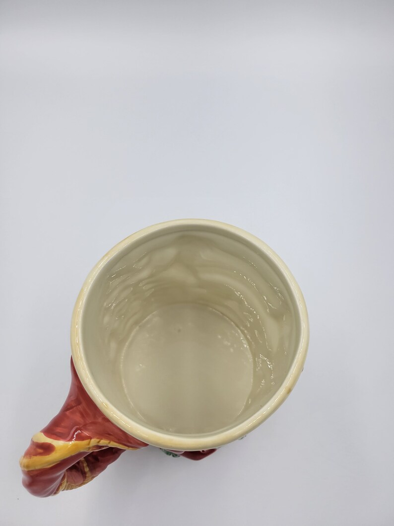 Pode incluir: Uma caneca de cer&acirc;mica com interior creme e uma al&ccedil;a vermelha com detalhes dourados e verdes. O interior da caneca tem uma superf&iacute;cie texturizada. A caneca est&aacute; sobre um fundo branco.