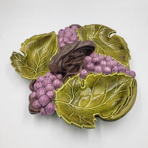 May include: A ceramic serving dish shaped like three green grape leaves arranged in a cluster. The dish features raised, glossy purple grape bunches and a central dark brown stem element. The surface has a smooth, glazed finish throughout.