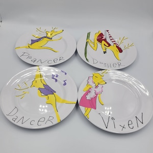 May include: Four white ceramic plates with hand-painted reindeer illustrations. Each plate features a different reindeer character: Prancer, Dasher, Dancer, and Vixen. The reindeer are yellow with various outfits and accessories. The names of the reindeer are written in black.