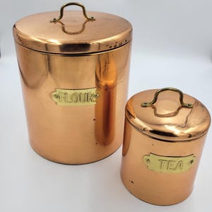 May include: Two cylindrical copper canisters with matching lids and brass handles. The larger canister features a brass label plate with the word FLOUR, while the smaller canister has a brass label plate with the word TEA, both in a metallic finish.