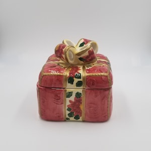 May include: A ceramic gift box with a bow on top. The box is red with gold accents and a floral pattern. The bow is cream-colored with red floral details. The box is rectangular and has a lid.