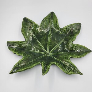 May include: A glossy, dark green ceramic leaf-shaped serving dish. The dish has a detailed vein pattern and a slightly raised edge. The overall shape is reminiscent of a large, stylized leaf, perfect for serving appetizers or as a decorative piece.