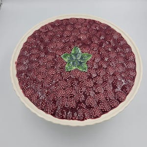May include: A round ceramic serving platter featuring a raised, textured surface molded to resemble a dense layer of dark red raspberries. A sculpted green leaf accent is positioned in the center of the fruit design, surrounded by a scalloped cream-colored rim.