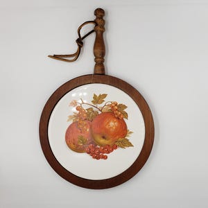 May include: A round ceramic trivet featuring a central illustration of red apples and currants with green leaves. The ceramic insert is framed by a dark wood border and includes a matching wooden handle at the top with a brown leather hanging strap.