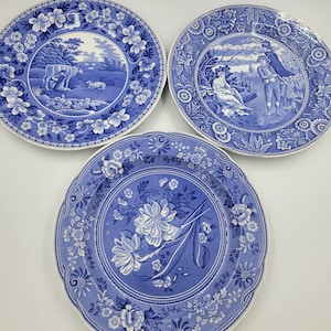 May include: Three blue and white ceramic plates with floral designs. The plates feature different scenes, including a pastoral scene with cows and sheep, a couple sitting under a tree, and a floral design.