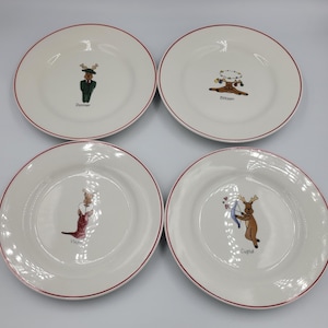 May include: Four white ceramic plates with red rims, each featuring a different cartoon reindeer character. The reindeer are named Donner, Blitzen, Vixen, and Cupid. The plates are likely for decorative or holiday use.
