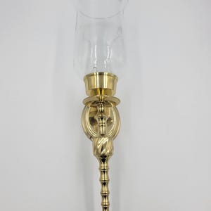 Pode incluir: Um candeeiro de parede para vela com uma base de metal dourado polido com um design vertical, cónico e canelado. O acessório inclui uma placa de montagem circular e um abajur cilíndrico de vidro transparente sobre o copo de metal.