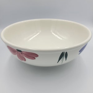 May include: Cream-coloured ceramic bowl with a floral pattern. The bowl has a wide rim and is decorated with pink, purple, and green painted flowers and leaves. The bowl is likely used for serving food.