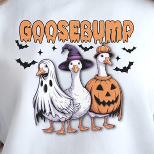May include: A white sweatshirt featuring a graphic of three geese in Halloween costumes. One goose wears a ghost sheet, one a purple witch hat, and one a pumpkin suit. The text GOOSEBUMP appears in orange above the geese with black bats.