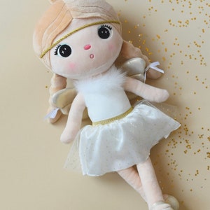 May include: A white plush angel doll with blonde hair, a gold headband, and a white tulle skirt with gold glitter. The doll has large, black eyes and a pink nose.