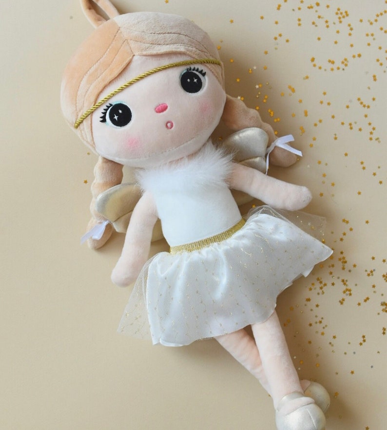 May include: A white plush angel doll with blonde hair, a gold headband, and a white tulle skirt with gold glitter. The doll has large, black eyes and a pink nose.