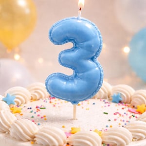 May include: A light blue number three birthday candle with a lit wick, positioned upright on a round cake decorated with white frosting swirls, colourful sprinkles, and small yellow, pink, and blue star-shaped sugar decorations.