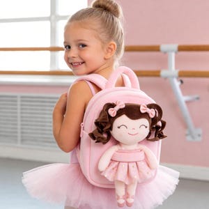 Puede incluir: Una mochila de color rosa claro suave con una muñeca de peluche unida al frente. La muñeca tiene cabello castaño en coletas con pequeños lazos rosas, una cara sonriente y viste un vestido rosa con estrellas doradas y zapatillas de ballet.