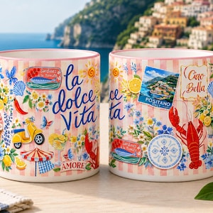 May include: Two ceramic mugs with pink handles and Italian-themed designs. The mugs feature a pink and white striped pattern, with illustrations of lemons, flowers, a scooter, and the phrases "la dolce Vita" and "Ciao Bella".