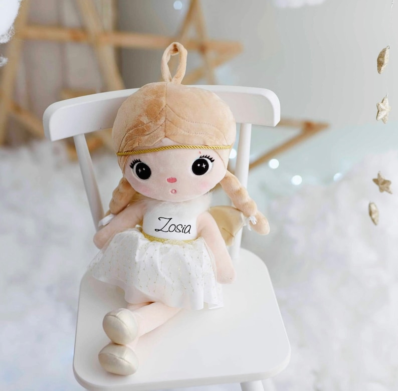 May include: A plush angel doll with blonde hair, a white dress, and gold wings. The doll is sitting on a white chair and has the name "Zosia" embroidered on its dress.