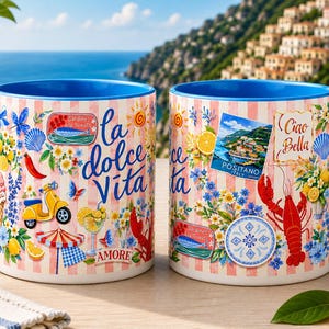May include: Two ceramic mugs with blue handles and rims, decorated with Italian-themed illustrations. One mug features the text "la dolce Vita" and the other "Ciao Bella". The mugs are adorned with images of lemons, a scooter, and a lobster.