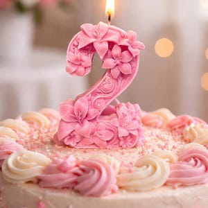 Può includere: Una candela rosa a forma di numero due con intagli floreali in rilievo e uno stoppino acceso sulla sommità. La candela è posizionata sopra una torta rotonda decorata con vortici di glassa bianca e rosa e piccoli zuccherini rosa.