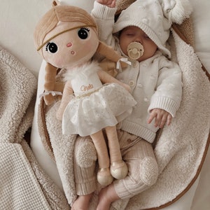 May include: A baby wearing a white knitted hat and sweater with a brown and white plush doll. The doll is wearing a white dress with gold glitter and the name "Nella" written on it. The baby is holding a pacifier in their mouth.
