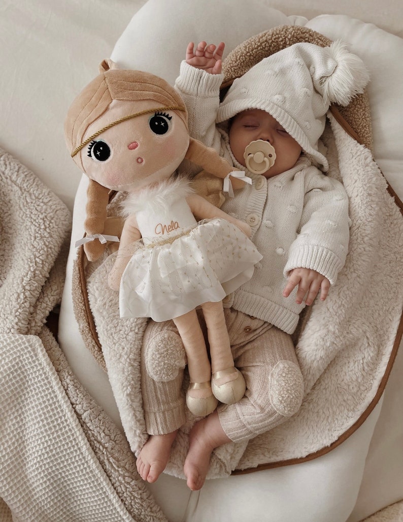 May include: A baby wearing a white knitted hat and sweater with a brown and white plush doll. The doll is wearing a white dress with gold glitter and the name "Nella" written on it. The baby is holding a pacifier in their mouth.