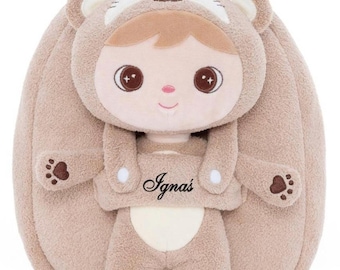 May include: A tan plush backpack featuring a soft doll character wearing a bear hood. The doll has large brown eyes, a small pink nose, and a cream-colored belly. The name Ignas is embroidered in black script on the doll's chest. The backpack has bear paw details on the doll's hands and feet.