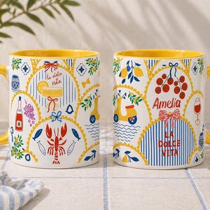 Puede incluir: Dos tazas de cer&aacute;mica blanca con asas e interiores amarillos. Las tazas presentan coloridas ilustraciones de tem&aacute;tica italiana, incluyendo limones, un scooter y las palabras "La Dolce Vita" y "Amelia".