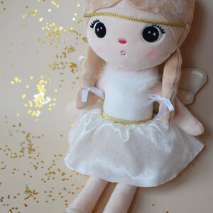 May include: A plush angel doll with blonde hair, a white dress with gold trim, and white wings. The doll is sitting on a light brown surface with gold glitter.