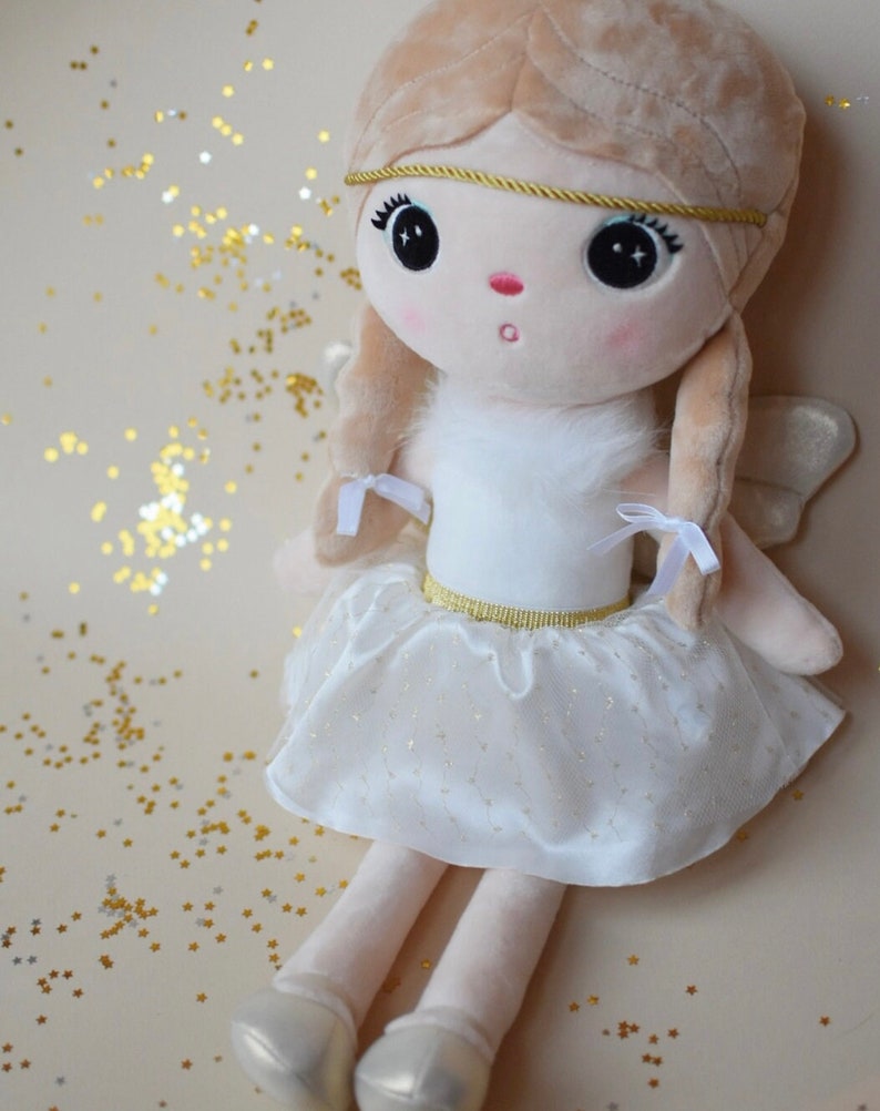 May include: A plush angel doll with blonde hair, a white dress with gold trim, and white wings. The doll is sitting on a light brown surface with gold glitter.