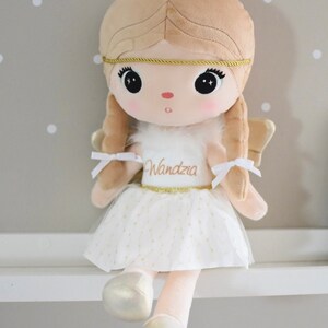 May include: A plush angel doll with blonde hair, a white dress with gold glitter, and gold wings. The doll is wearing a gold headband and has the name "Wandra" embroidered on her dress.