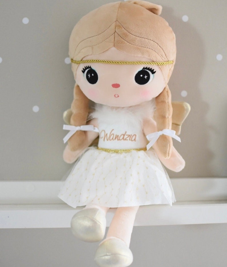 May include: A plush angel doll with blonde hair, a white dress with gold glitter, and gold wings. The doll is wearing a gold headband and has the name "Wandra" embroidered on her dress.