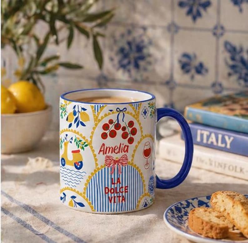 Puede incluir: Taza de cer&aacute;mica blanca con asa y borde azules, decorada con ilustraciones de tem&aacute;tica italiana. La taza presenta un scooter, limones, cerezas y el nombre "Amelia" con el texto "La Dolce Vita".