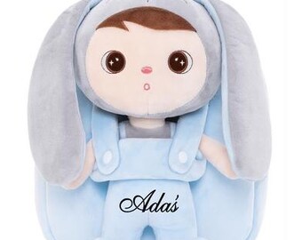 May include: A light blue plush backpack featuring a character wearing a grey bunny hood with long ears. The character has brown hair, large dark eyes, and is wearing light blue overalls with the name Ada's embroidered in black script on the front.