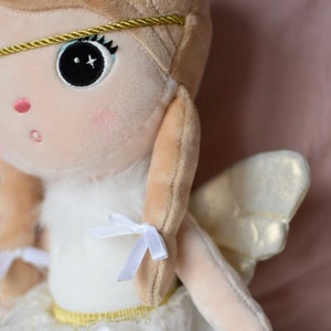 May include: A plush angel doll with blonde hair, a white dress, and gold wings. The doll has a sweet expression and is wearing a gold headband.