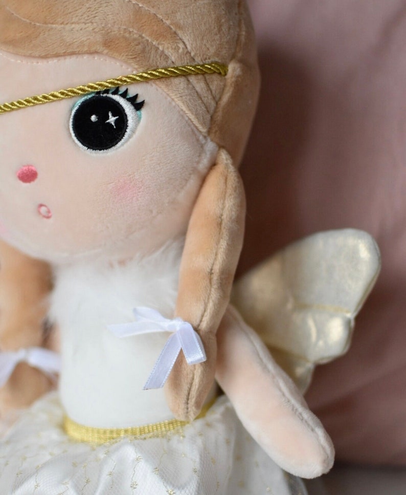 May include: A plush angel doll with blonde hair, a white dress, and gold wings. The doll has a sweet expression and is wearing a gold headband.