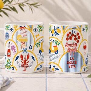 Puede incluir: Dos tazas de cer&aacute;mica blanca con dise&ntilde;os italianos coloridos. Una taza presenta una langosta, peces y el texto "la dolce vita". La otra taza tiene un scooter, cerezas y el nombre "Amelia". Ambas tazas tienen un asa curva.