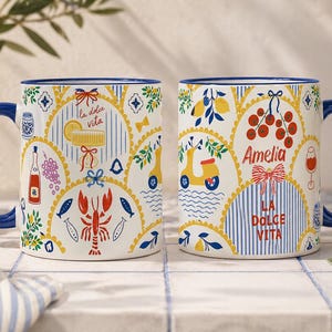 Puede incluir: Dos tazas de cer&aacute;mica blanca con asas y bordes azules. Las tazas presentan ilustraciones coloridas de limones, un scooter y el texto "La Dolce Vita". Una taza tiene el nombre "Amelia".