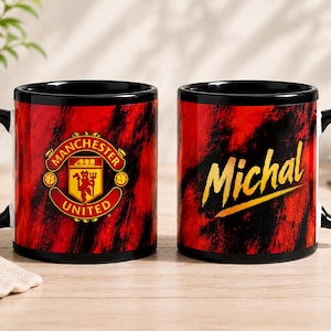 May include: Two ceramic mugs with a black handle and rim, featuring a red and black brushstroke pattern. One mug displays the Manchester United logo in red, yellow, and black. The other mug features the name Michal in yellow cursive script.