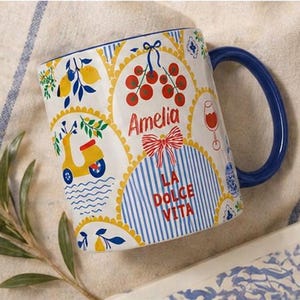 Puede incluir: Taza de cer&aacute;mica blanca con asa azul, decorada con ilustraciones de tem&aacute;tica italiana. Presenta un scooter amarillo, limones y el texto "Amelia" y "La Dolce Vita" en rojo. El dise&ntilde;o incluye rayas azules y blancas y un lazo rojo.