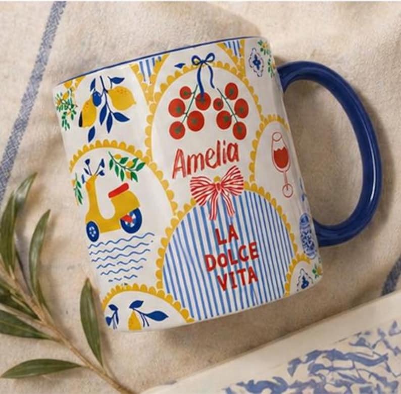 Puede incluir: Taza de cer&aacute;mica blanca con asa azul, decorada con ilustraciones de tem&aacute;tica italiana. Presenta un scooter amarillo, limones y el texto "Amelia" y "La Dolce Vita" en rojo. El dise&ntilde;o incluye rayas azules y blancas y un lazo rojo.