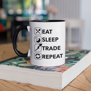 May include: White ceramic mug with a black interior and handle. The mug features the words "EAT SLEEP TRADE REPEAT" in black, with corresponding icons. The mug is sitting on a book with a floral design.