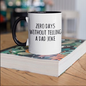 May include: White ceramic mug with a black handle and rim, featuring the text "ZERO DAYS WITHOUT TELLING A DAD JOKE" in black. The mug sits on a book with a colorful floral design, placed on a wooden surface.