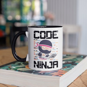 Puede incluir: Taza de cerámica blanca con asa y borde negros. La taza tiene un gráfico de un ninja de dibujos animados sosteniendo una computadora portátil con el texto "CODE NINJA" en negro.