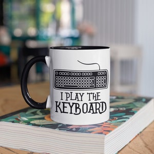 Może przedstawiać: Biały kubek ceramiczny z czarnym rantem i uchwytem. Kubek ma czarno-białą ilustrację klawiatury komputerowej z tekstem "I play the keyboard".