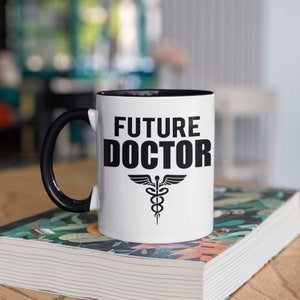 Può includere: Tazza in ceramica bianca con interno e manico neri. La tazza presenta la scritta "FUTURE DOCTOR" in grassetto nero, insieme a un simbolo di caduceo nero. La tazza è appoggiata su un libro.