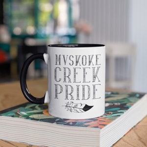 May include: White ceramic mug with a black rim and handle. The mug has black text that reads "MVSKOKE CREEK PRIDE" with a stylized feather below the text.