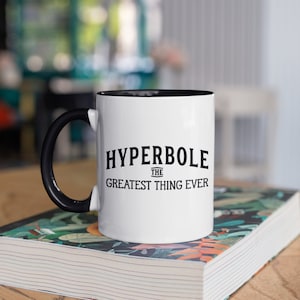 Puede incluir: Taza de cerámica blanca con asa e interior negros. La taza presenta el texto "HYPERBOLE THE GREATEST THING EVER" en negro, centrado en la parte delantera. La taza está sobre un libro con diseño floral.