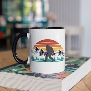 May include: White ceramic mug with a black handle and interior. The mug features a graphic of a Bigfoot silhouette walking in front of a sunset with mountains and trees. The mug is sitting on a book.