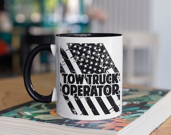 May include: White ceramic mug with a black handle and interior. The mug features a distressed American flag design with the words "TOW TRUCK OPERATOR" in bold, black letters. The mug is sitting on a book with a floral design.