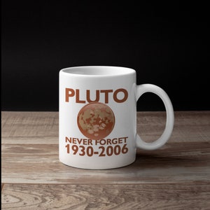 Peut inclure: Mug en céramique blanche avec le mot "PLUTO" en lettres marron au-dessus d'un dessin marron et beige de Pluton. Le texte "NEVER FORGET 1930-2006" est en dessous. La tasse a une anse incurvée.