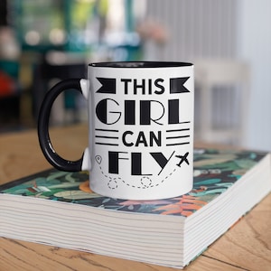 May include: White ceramic mug with a black rim and handle. The mug has a black graphic that says "This Girl Can Fly" with a plane and a dotted line.