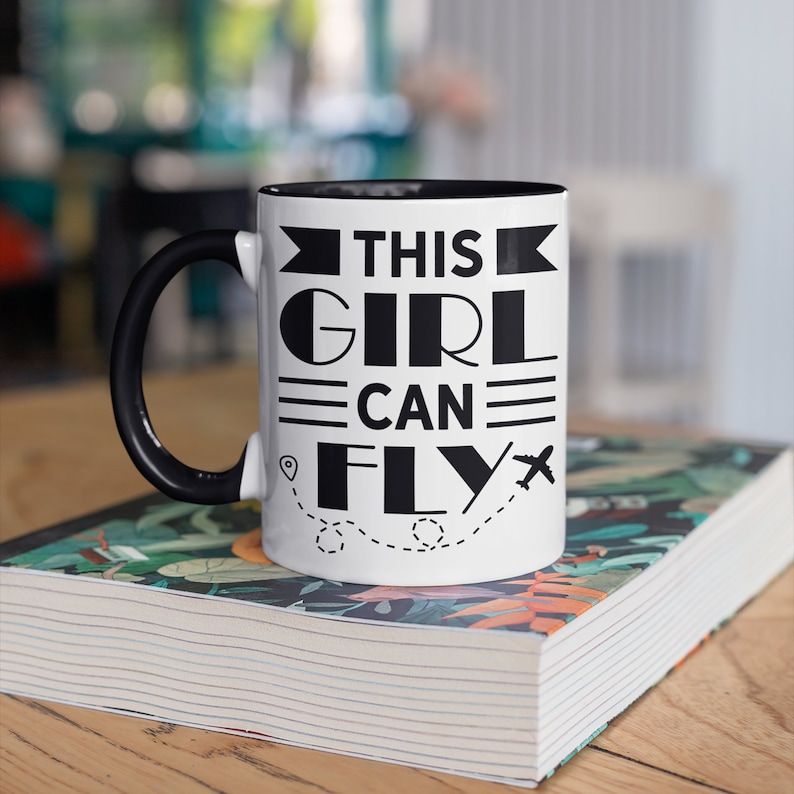 May include: White ceramic mug with a black rim and handle. The mug has a black graphic that says "This Girl Can Fly" with a plane and a dotted line.